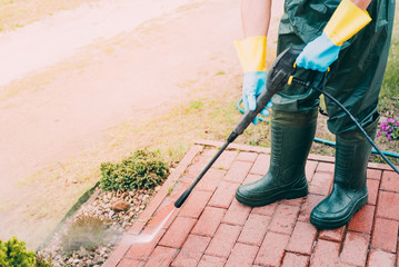 Man cleaning red, conrete pavement block using high pressure water cleaner. Paving cleaning concept. Man wearing waders, rubber boots, gloves and waterproof trousers doing spring jobs in the garden