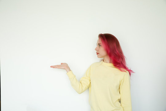 Photo of a young woman wearing yellow shirt, standing isolated over white wall background. Pointing to copy space, neutral palette. 