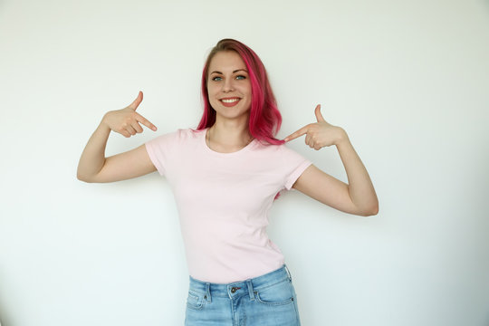 Photo of a very happy young woman standing isolated over white wall background. Pointing to copy space in a pink shirt, neutral palette.