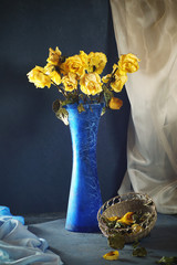 A composition of yellow roses standing in a blue, glass vase, next to an openwork fruit bowl filled...