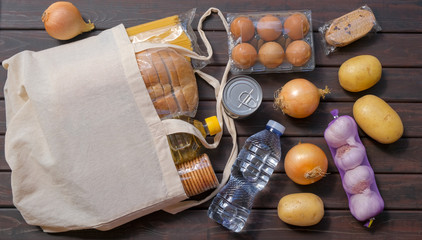 Textile bag with different food on dark wooden background. Food supplies, bread, fruit, vegetables, pasta, eggs, canned food, cookies. Food stock. Quarantine, isolation. Safe home delivery.