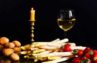 Uncooked asparagus, potatos and strawberries in front of a black background, with a glass of wine, a jar and a candle, like a still life, like a painting of an ancient artist