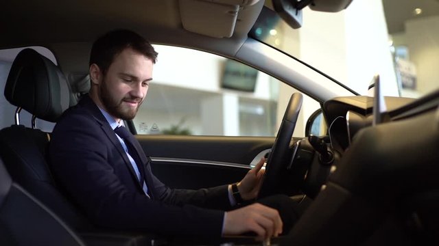 Stylish businessman wearing suit gets behind the wheel of car he bought in auto dealership, view from interior of car. Concept of choosing and buying new car at showroom. Shooting in slow motion.