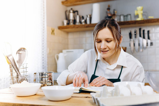 Close Up Image Of Woman Sitting At Wooden Kitchen Table Looking To  Recipe Book And Trying To Choose What To Cook. Cooking At Home Concept, Lifestyle. Ketogenic Diet And Menu