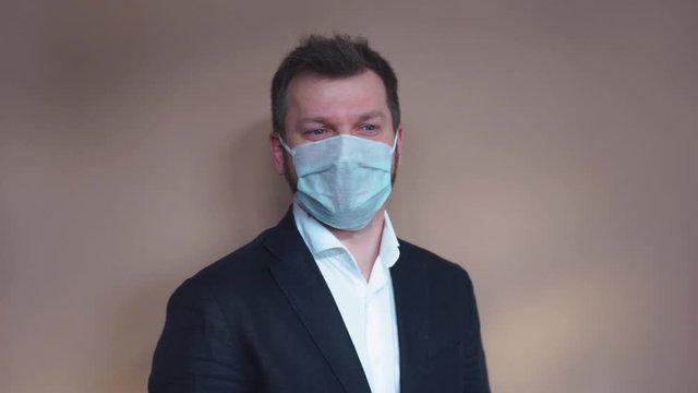 Handsome man in a white T-shirt puts on a medical protective mask and looks at the camera against a beige background. Portrait, the average plan. Natural light, soft focus.