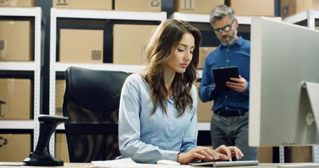 Beautiful female postal worker working at computer in post delivery office and typing on keyboard. Young woman in mail store sitting at table in storage.