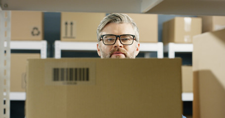 Close up portrait of caucasian handsome postman in glasses taking carton box with bar code from...