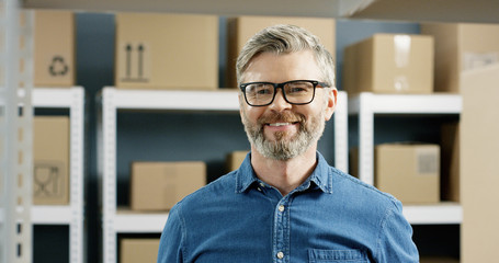 Close up portrait of happy smile postman in glasses standing in postal store with carton boxes on shelves. Post worker smiling joyfully to camera in mail office among parcels.