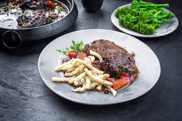 Traditional German braised beef cheeks in brown red wine sauce with noodles and broccoli offered as closeup on a modern design plate and stewpot on an old rustic board