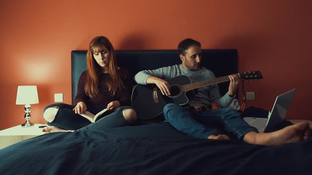 joven pareja sentados en la cama leyendo y tocando la guitarra