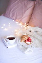 Fresh breakfast with cup of black tea on open paper book with croissant, ripe strawberry, whipped cream on white plate and knitted cloth in bed closeup over glowing lights. Good morning. Breakfast.