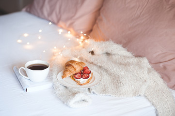 Fresh croissant with ripw strawberry and whipped cream on white plate, cup of black tea staying on paper book in bed closeup. Good morning.