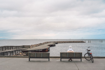 Fototapeta na wymiar A young Caucasian woman sits with her back on a wooden bench overlooking the Baltic Sea on the seafront in Copenhagen Denmark in winter in cloudy weather. Girl walking gonoskoy bike parked nearby