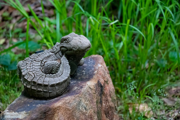 A lifelike alligator water sprinkler perches on a rock in a rustic garden against a background of tall grass. Concept of garden humor.