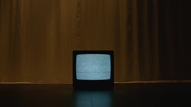 Old CRT TV is on the floor in a dark and empty room. On the screen there is a frightening white noise filling the room with a hypnotizing atmosphere of mystical evil. Dolly camera movement
