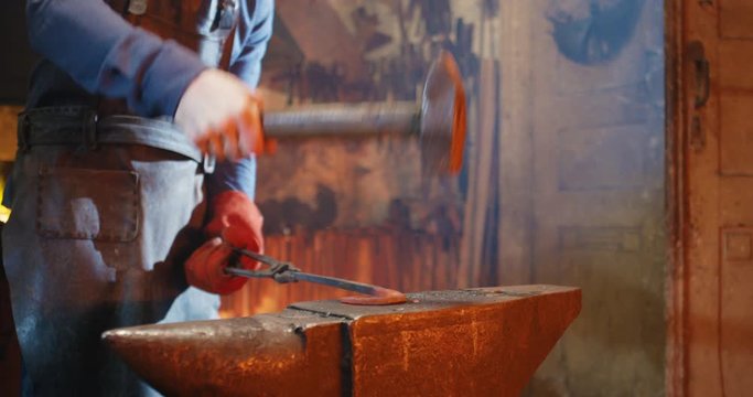 Close up of glowing metal item in hands of blacksmith whie he shaping it with hammer in smithy. Hard work of smith in forge. Metal antique manufactoring concept.