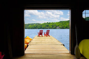 View of two red Adirondack chair on a wooden dock from a cottage's boathouse in Muskoka, Ontario Canada. In the background cottages are nestled between trees.