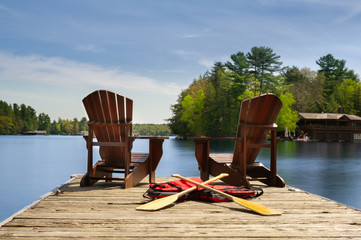 Two Adirondack chairs on a wooden dock facing the blue water of a lake in Muskoka, Ontario Canada....