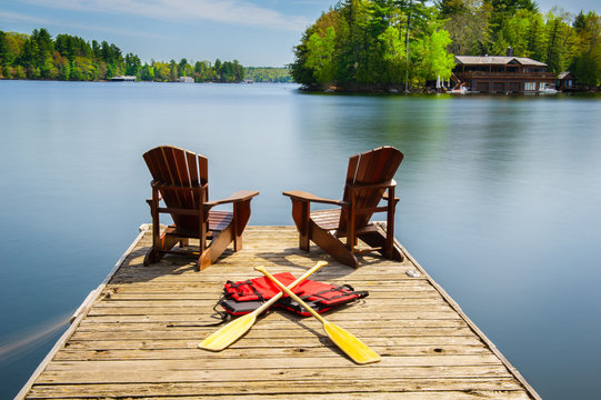 Two Adirondack chairs on a wooden dock facing the blue water of a lake in Muskoka, Ontario Canada. Canoe paddles and life jackets are on the dock. A cottage nestled between green trees is visible.