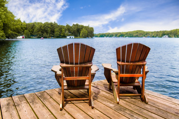 Two Adirondack chairs on a wooden dock overlooking a calm lake. Cottages nestled between green...
