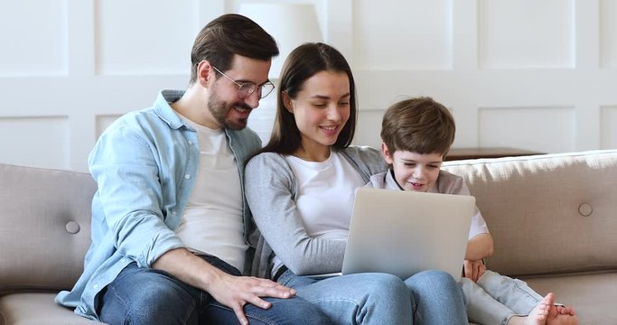 Happy loving family couple relaxing on sofa with little preschool child son, using computer, shopping in internet store at home. Smiling parents playing games online with small kid boy in living room.