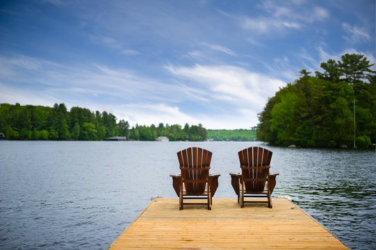 Two Adirondack chairs on a wooden dock overlooking a calm lake. Cottages nestled between green trees are visible across the water.