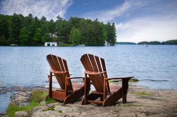 Fototapeta premium Two Muskoka chairs along the shore facing a lake in Ontario, Canada. A white cottage is visible across the blue water nestled between green trees.