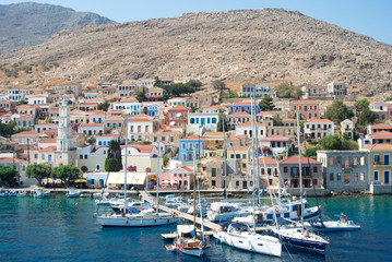 Fototapeta na wymiar The colourful harbour at the the beautiful Greek island of Chalki. Classical architecture rises from the waterfront looking over the bay. A warm summers day with pleasure boats moored. 