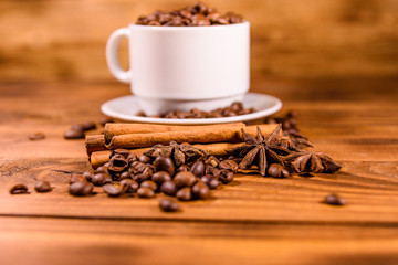 White cup filled with coffee beans, star anise and cinnamon sticks on wooden table