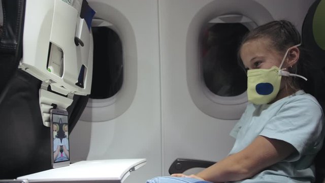 Little Girl Caucasian At Plane With Wearing Protective Medical Mask. Child Baby Tourist At Aircraft With Respirator Play At Smartphone Mobile Phone. Coronavirus Epidemic Sars-cov-2 Covid-19 2019-ncov.