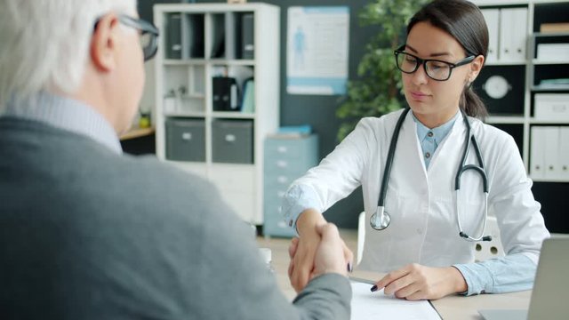 Young woman doctor is shaking hands with elderly man patient talking in office during individual consultation. People and medical help concept.