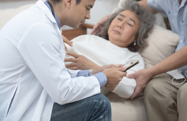 Fototapeta premium Asian senior elderly old patient woman laying down on sofa in home waiting for result from smart young caucasian doctor smartphone while check up in home with grandfather take care beside.