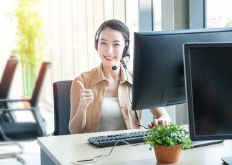 Attractive and smile young asian customer servicer woman wearing headset in modern creative meeting working office sit at computer monitor thump up ,work from and social distancing concept.