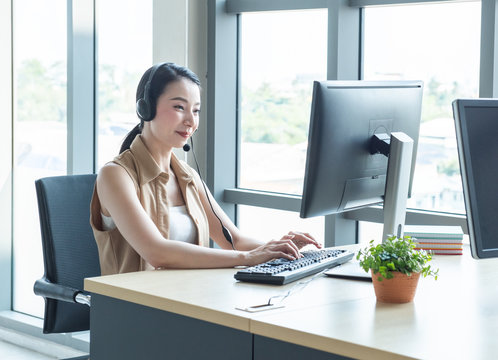 Attractive And Smile Young Asian Customer Servicer Woman Wearing Headset In Modern Creative Meeting Working Office Look At Computer Monitor,work From And Social Distancing Concept.