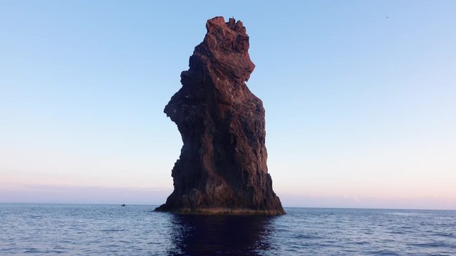 Rock in Mediterranean sea against horizon and Lipari Island. Colorful sky, sunrise. Rippling water surface. Sicily, Italy