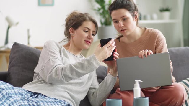 Young cheerful woman showing funny picture on smartphone to female friend, they discussing it and laughing while resting together on couch at home during quarantine