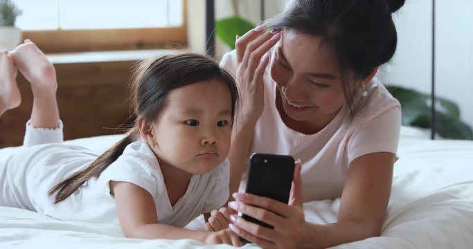 Cute little asian ethnicity preschool child girl lying on comfortable bed with young mommy nanny, using funny mobile applications. Happy vietnamese family having fun, recording video on cellphone.