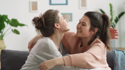 Two young Caucasian women taking medical face masks off, embracing each other and happily smiling while sitting together on couch at home and celebrating the end of coronavirus lockdown - Powered by Adobe