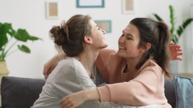 Two young Caucasian women taking medical face masks off, embracing each other and happily smiling while sitting together on couch at home and celebrating the end of coronavirus lockdown