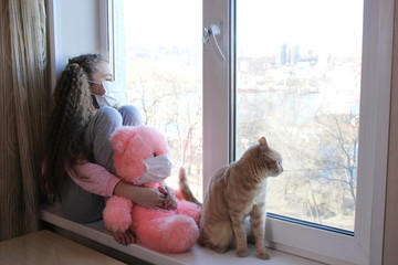 A girl in a protective medical mask on her face sits at home on the windowsill and looks out the...