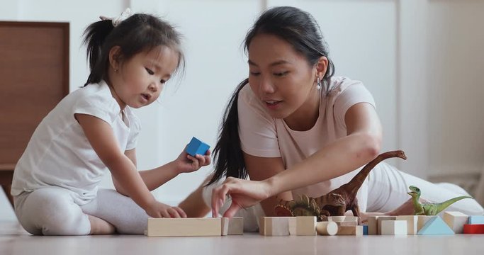 Excited young vietnamese nanny babysitter lying on heated floor with cute preschool child girl, playing with favorite toys at home. Happy asian mommy enjoying free leisure time with baby daughter.