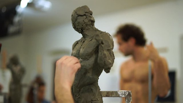 Student working on clay sculpture, nude model in background