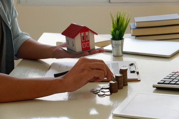 Saving money concept preset by Male hand putting money coin and signs a contract to purchase a home with a real estate agent. Model house with keys on table.