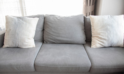 Close-up picture of grey soft with side pillows are comfortable, yet stylish and modern, for the whole family to spend their free time resting under the evening light that pass through the window.