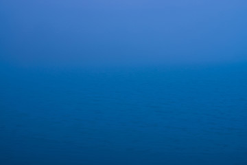 Fototapeta na wymiar Natural texture of deep blue calm water in dusk close up. Night sea of blue classic color. Water ripple nature background. Meditative image of thick fog above lake. Soft light shines on water surface.