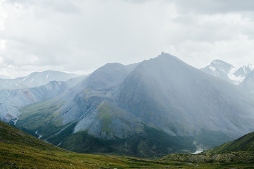 Fototapeta premium Atmospheric alpine view from pass to great mountain with sharp pinnacle under gloomy cloudy sky. Wonderful giant pointy rocky top and snowy mountains behind mountain pass. Awesome beauty of highlands.