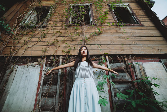 Crazy girl spread her arms like wings. Beautiful mad country girl near old rustic house with broken windows among summer greenery. Unusual female portrait on background of wooden abandoned aged house.