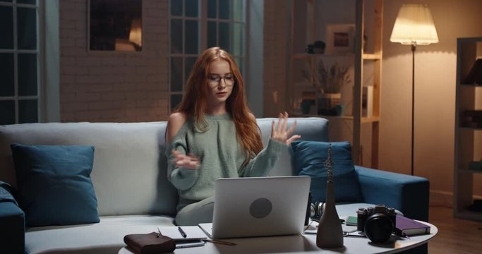 Pretty caucasian redhead girl sitting on coach looking at laptop, having a videochat or online tutorial, recording an internet vlog, cute model looking at webcamera and talking 4k footage