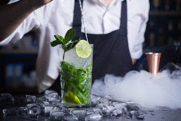 Glass of mojito dressed up with lemon and mint. Ice cubes placed on surface around mojito drink. Little white cloud of smoke hiding measuring cup.Barmen in white top and black apron stirring cocktail - obrazy, fototapety, plakaty