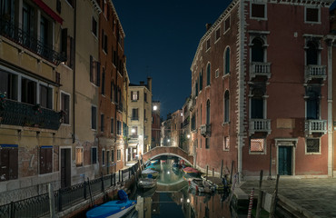 A scenic view of a beautiful Venetian canal at night with colourful architecture, a bridge and reflections of boats running along the water in the town of Venice, Italy